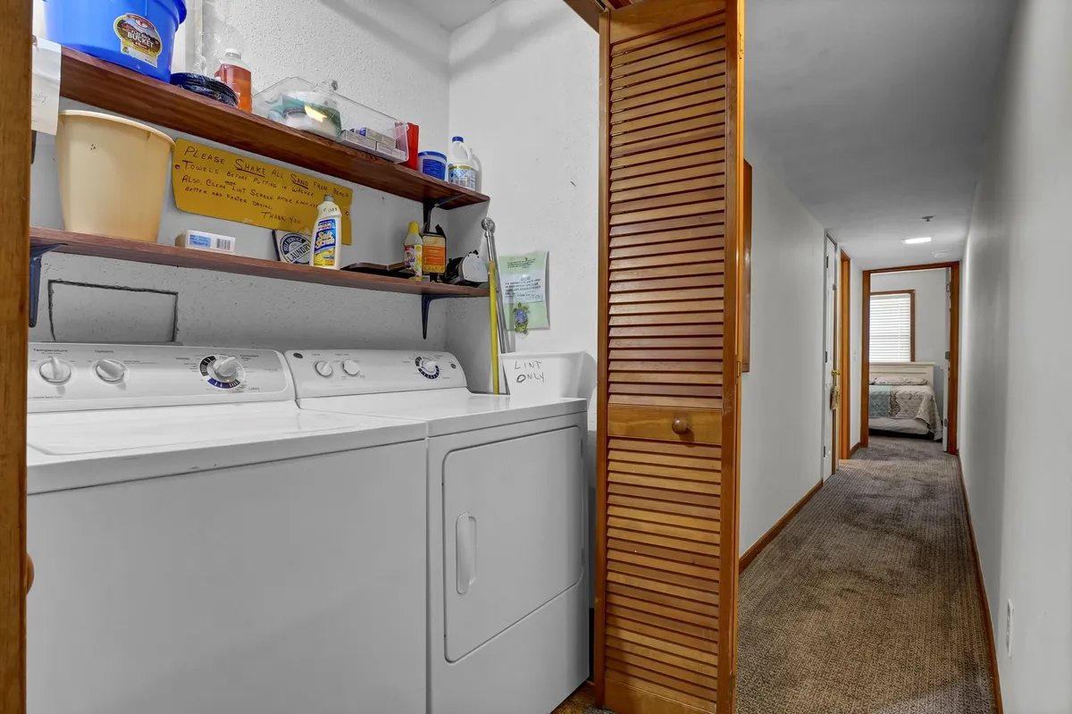 A practical laundry area featuring a washing machine and dryer, shelves stocked with laundry supplies, and a view into the hallway, emphasizing convenience and organization.