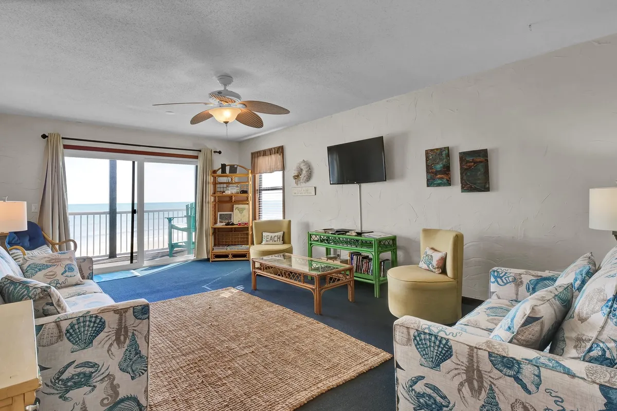 The image showcases a spacious living room with comfortable seating arranged to face large windows that offer stunning ocean views. The decor includes colorful accents and a glass coffee table with a beach-inspired theme, creating a cheerful and relaxed atmosphere. The space invites socializing, reading, and unwinding while making the most of the spectacular coastal setting.