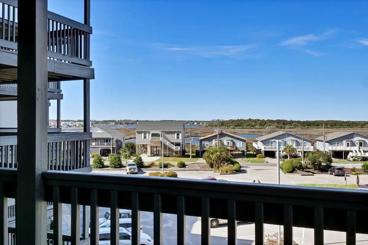 This image showcases a serene waterfront view with surrounding houses, lush greenery, and a glimpse of a parking area. The vantage point appears to be from a private balcony, inviting viewers to imagine themselves relaxing while taking in the tranquil coastal atmosphere.