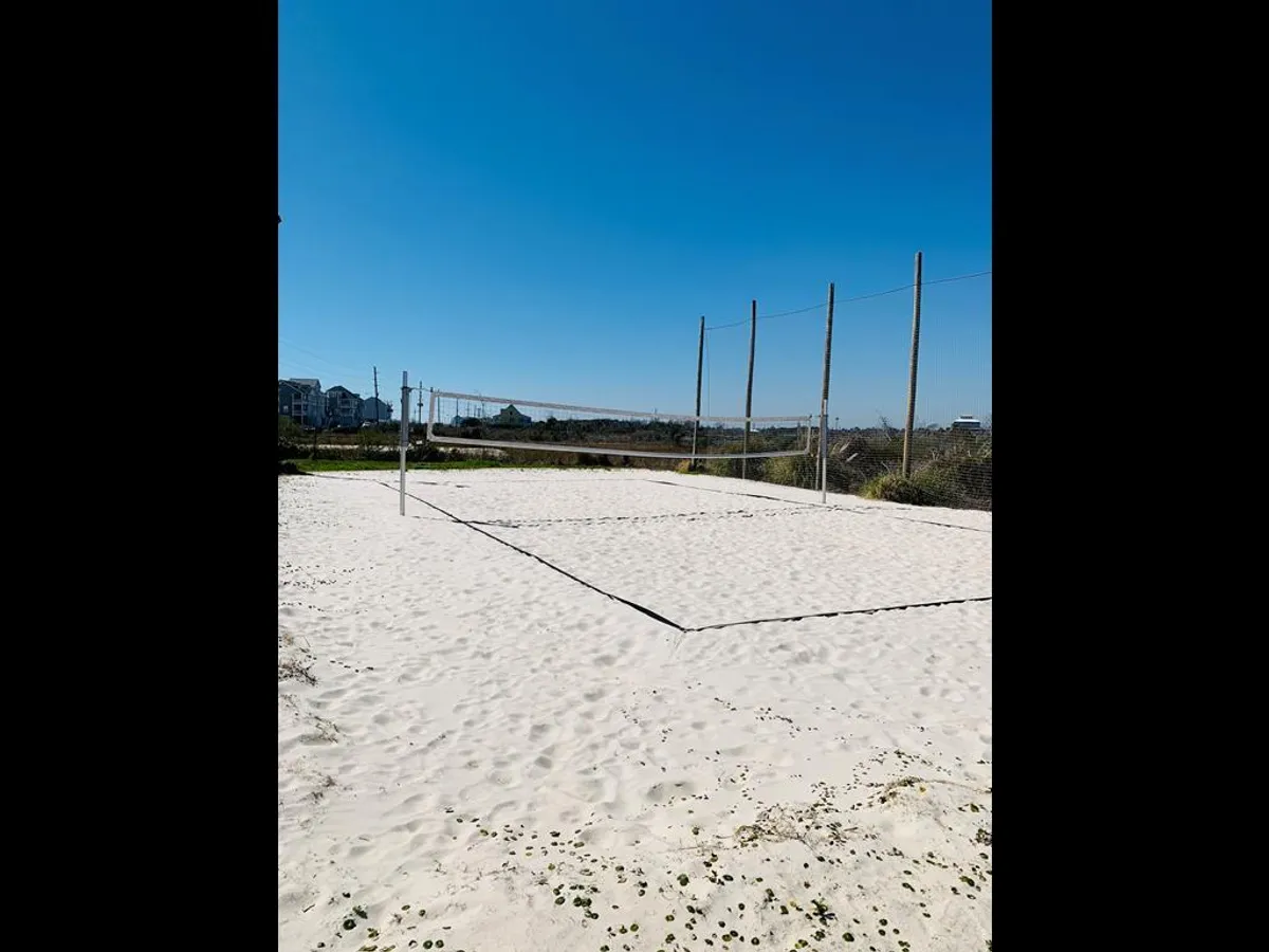 A pristine white sand volleyball court sits against a backdrop of blue sky, with ample space for beach games and group activities. The area is lively and energetic, perfectly suited for friendly matches or spontaneous games with friends.
