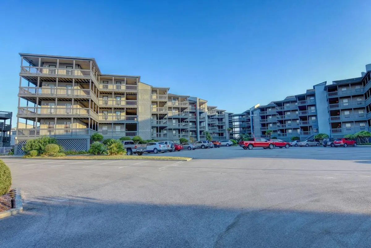 This exterior image features the multi-story Shipwatch condominium buildings surrounded by landscaped greenery and a parking area. Balconies are visible, suggesting inviting spaces for relaxing or working with fresh air and an ocean breeze nearby.