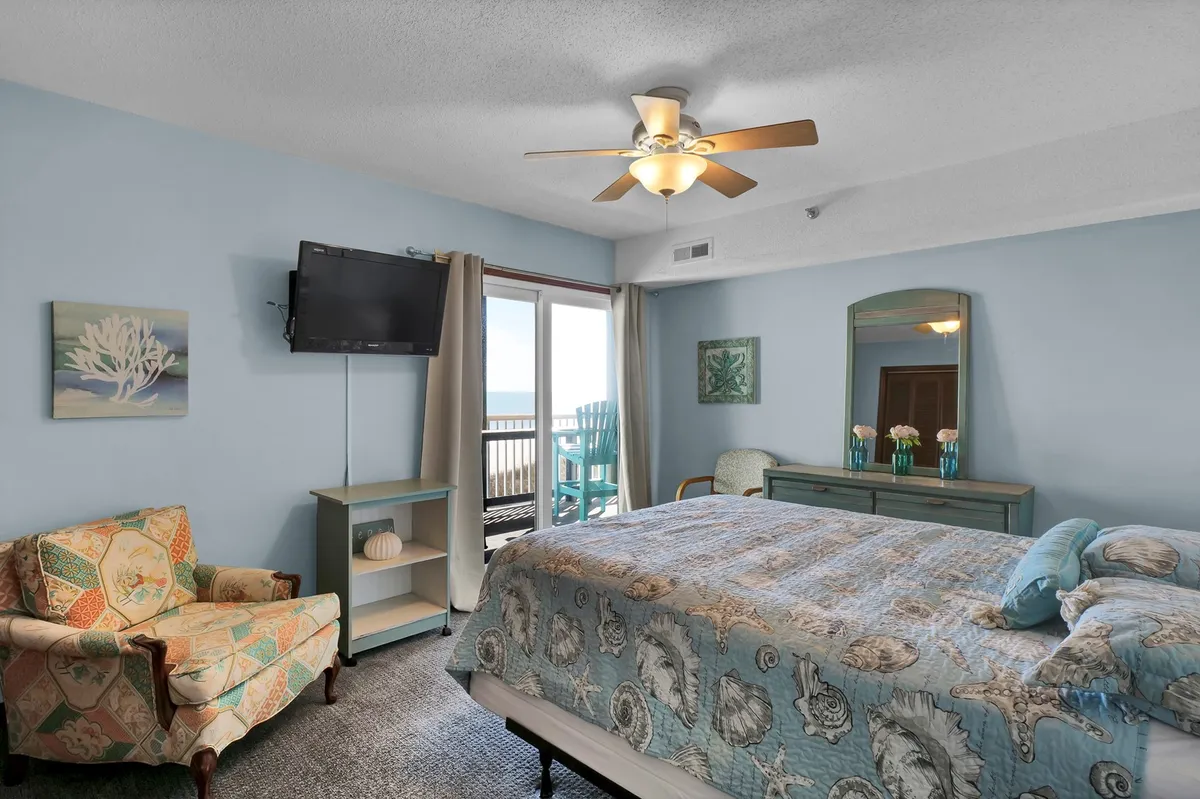 This image displays a tranquil master bedroom, featuring a plush bed positioned near a glass balcony door. An armchair in the corner creates a cozy reading nook, while a dresser and a television add both comfort and practicality. Soft natural light enters through the balcony door, hinting at the private outdoor space accessible from this suite.