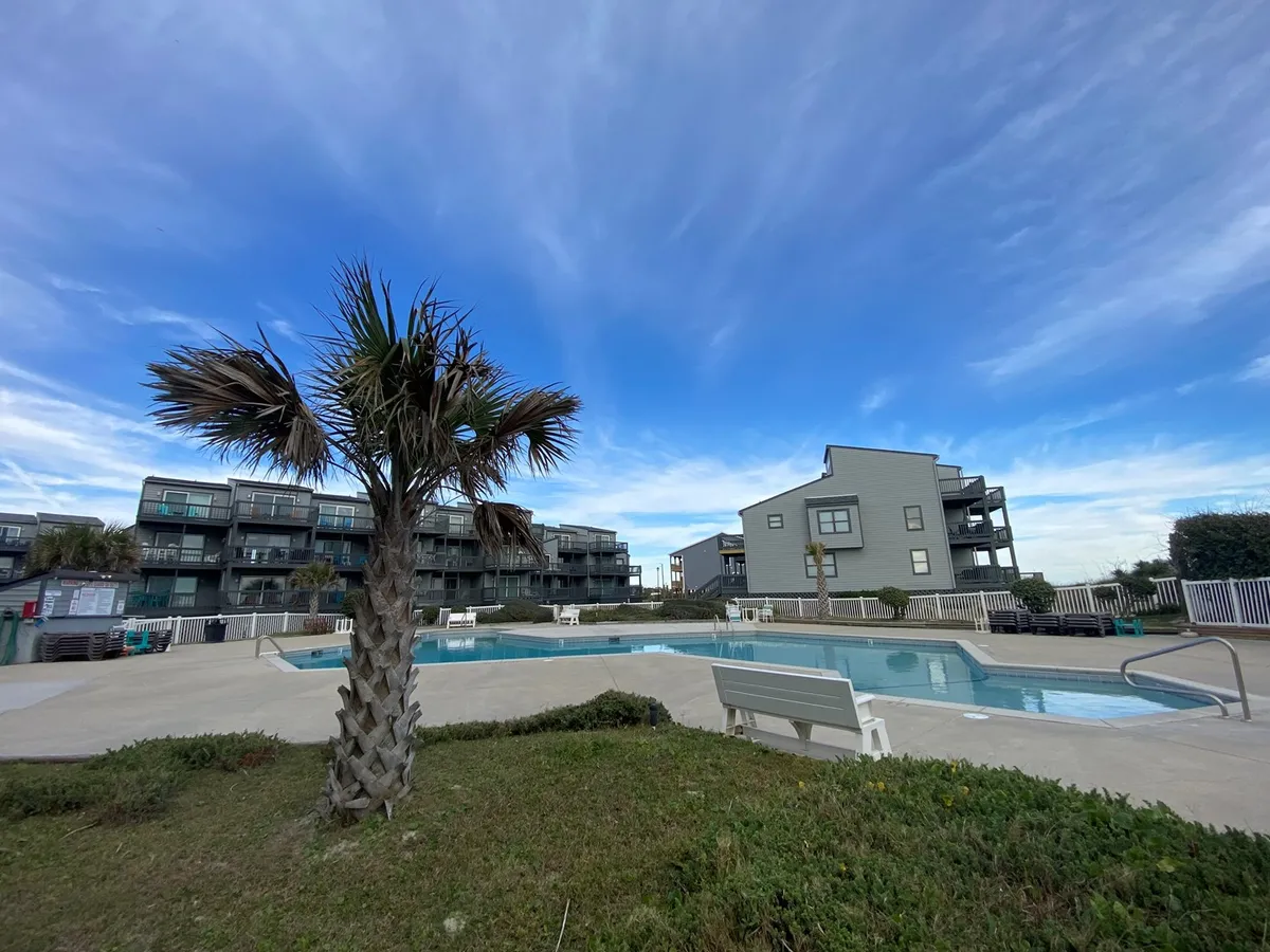 A tranquil scene shows a sparkling swimming pool surrounded by palm trees and modern condominium buildings. The mood is relaxed, offering spaces for swimming, sunbathing, and unwinding by the pool under the open sky.