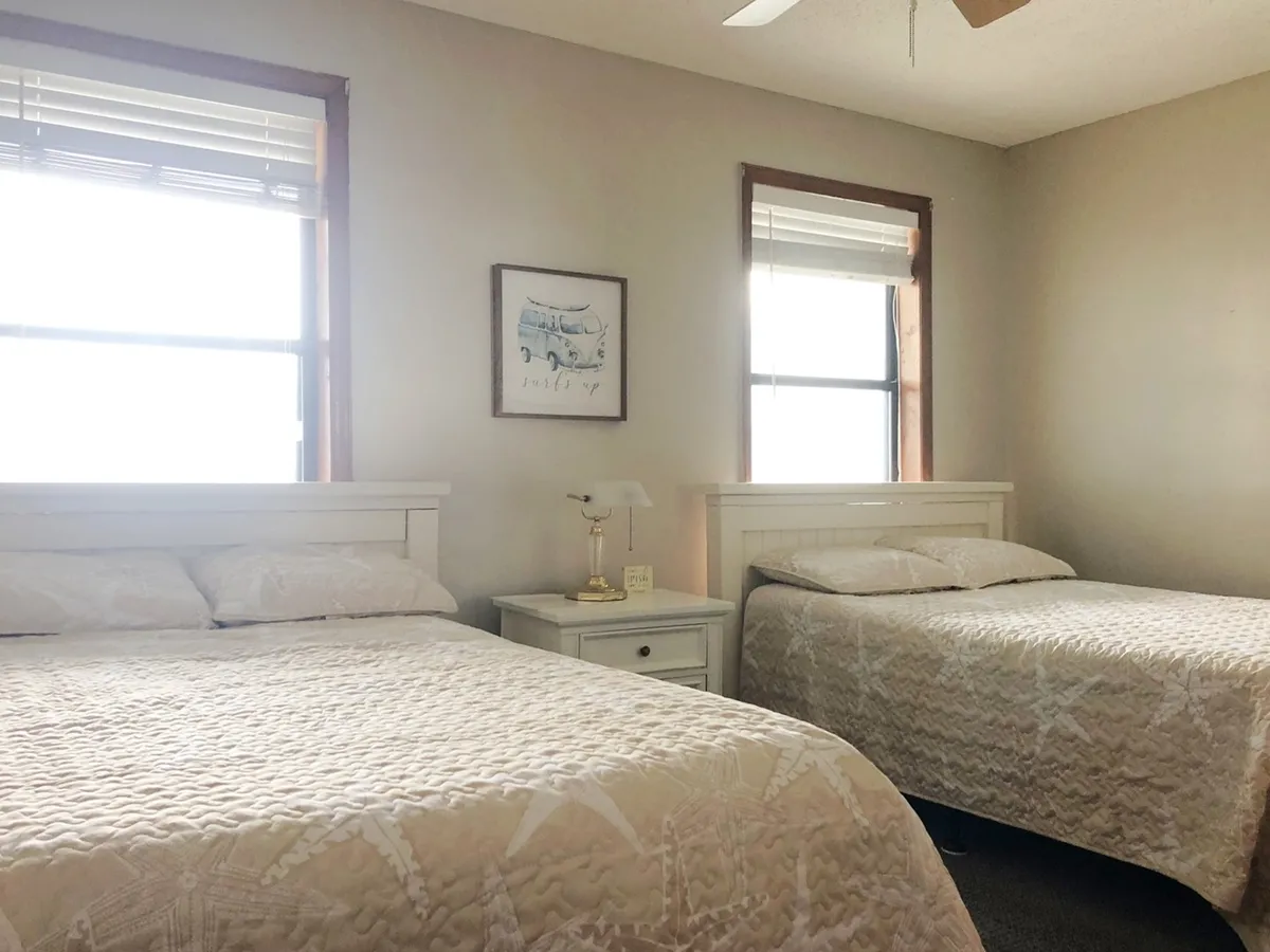 This photograph presents the second bedroom, showcasing two full-sized beds with crisp bedding. A nightstand sits between the beds, topped with a lamp, and large windows allow sunlight to brighten the room. The decor is soothing, with soft colors and tasteful wall art.