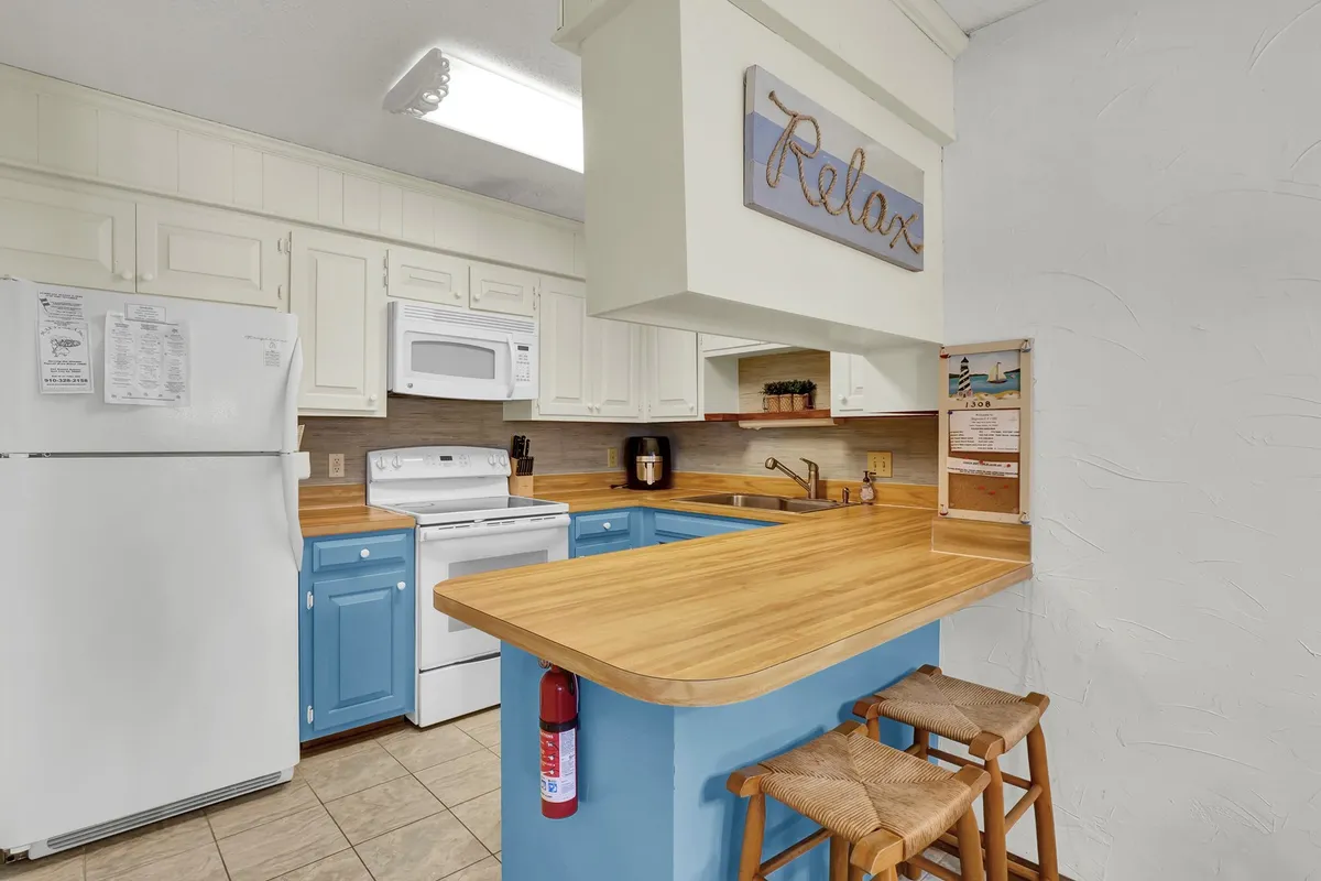 Featuring a modern kitchen with blue cabinetry, a wooden countertop, and bar stools at a breakfast bar, this image emphasizes the casual, inviting atmosphere for morning coffee, snacks, or casual dining. The 'Relax' sign adds a playful, coastal touch, underscoring the easy-living theme.